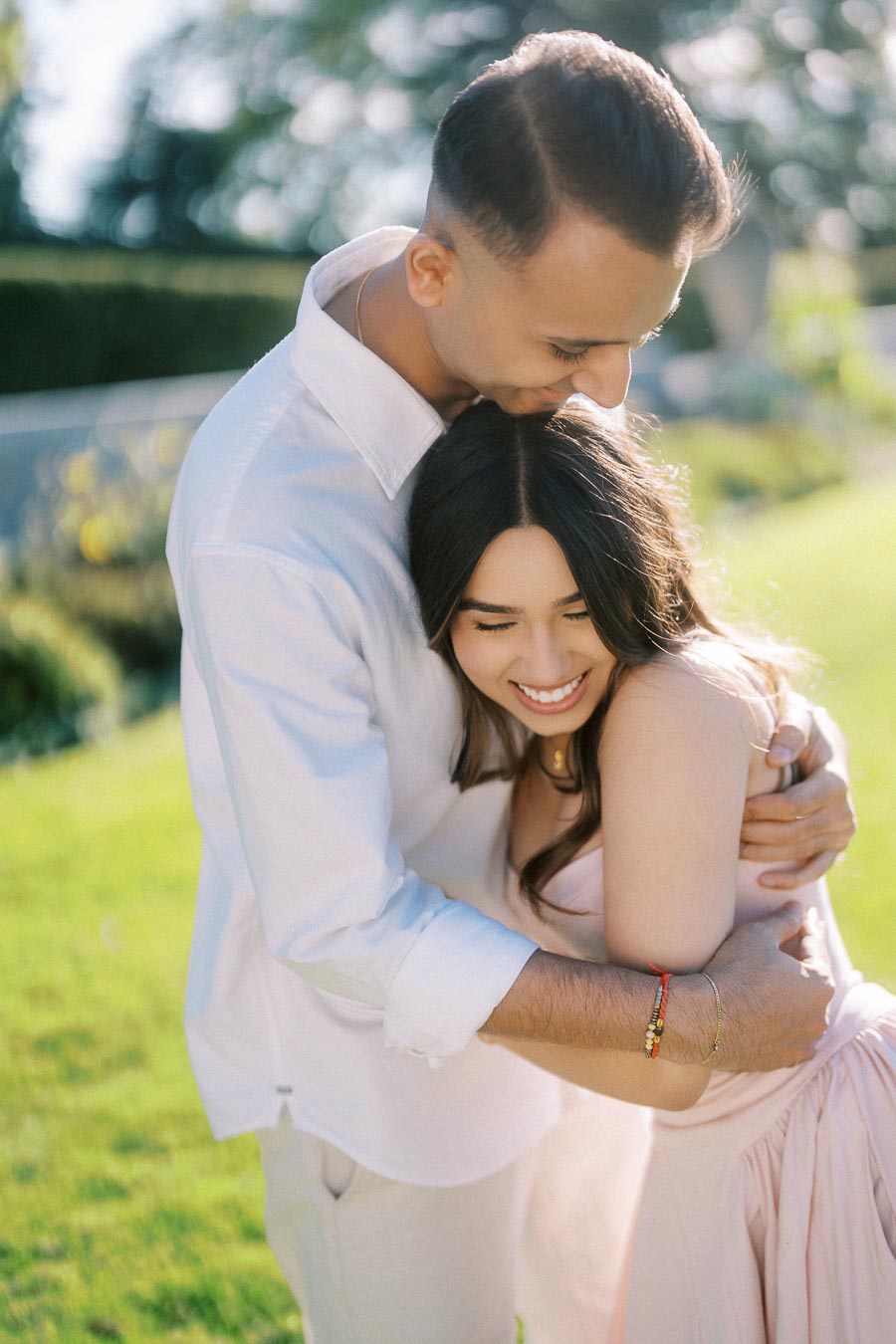 A couple embraces outdoors on a sunny day, with greenery in the background; the man kisses the woman's forehead as she smiles, wearing light, casual clothing.