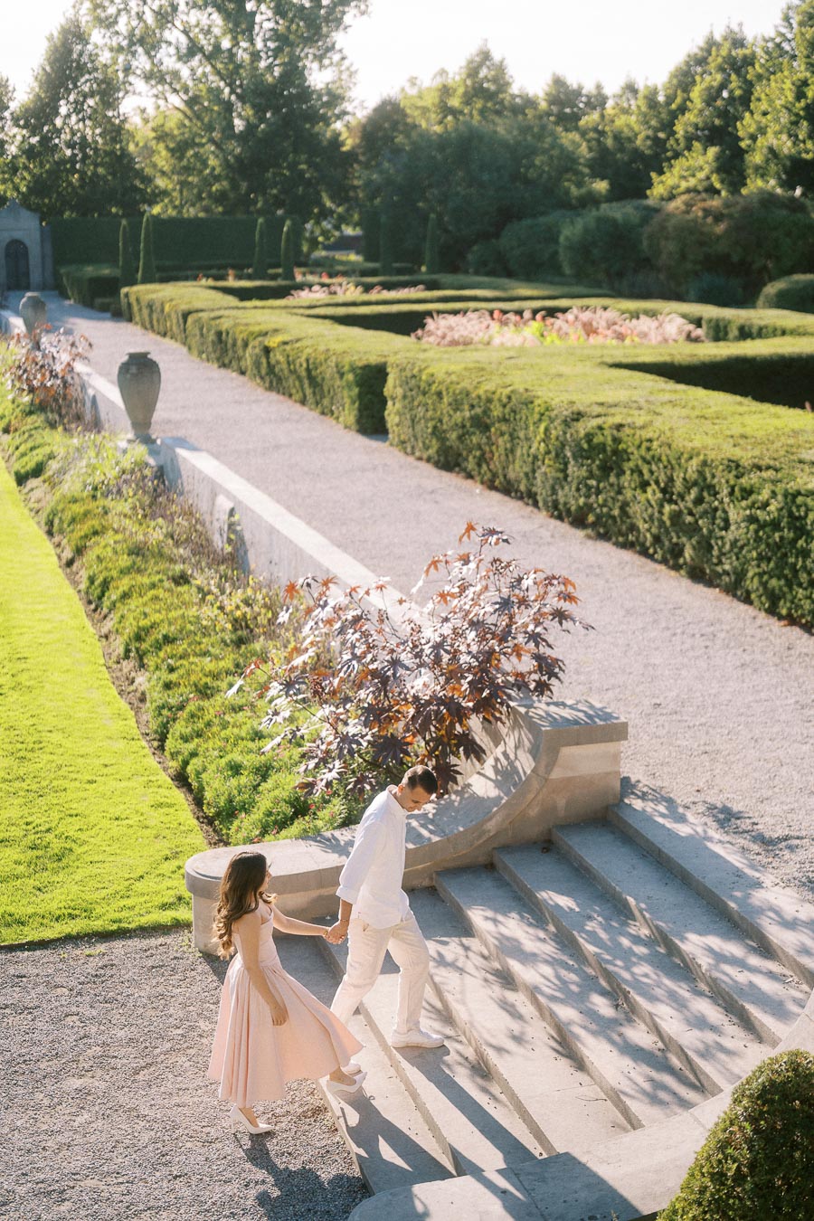 A couple holding hands and walking down stone steps in a beautifully landscaped garden, surrounded by vibrant green hedges and trees under a sunny sky.
