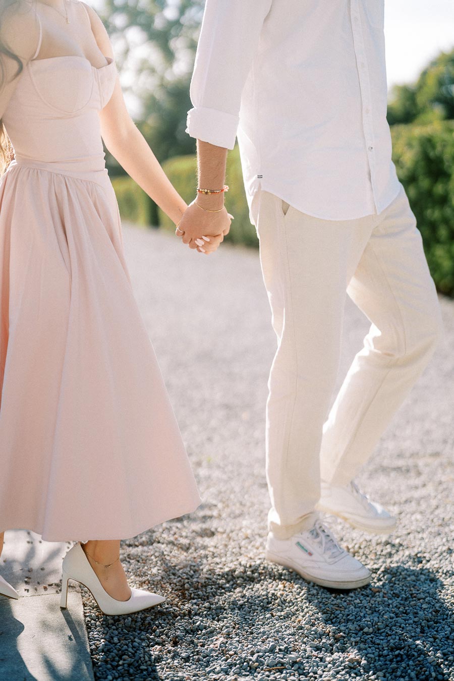 Elegant couple holding hands, walking together on a sunny day, with the woman in a light pink dress and heels, and the man in a white shirt and beige pants.
