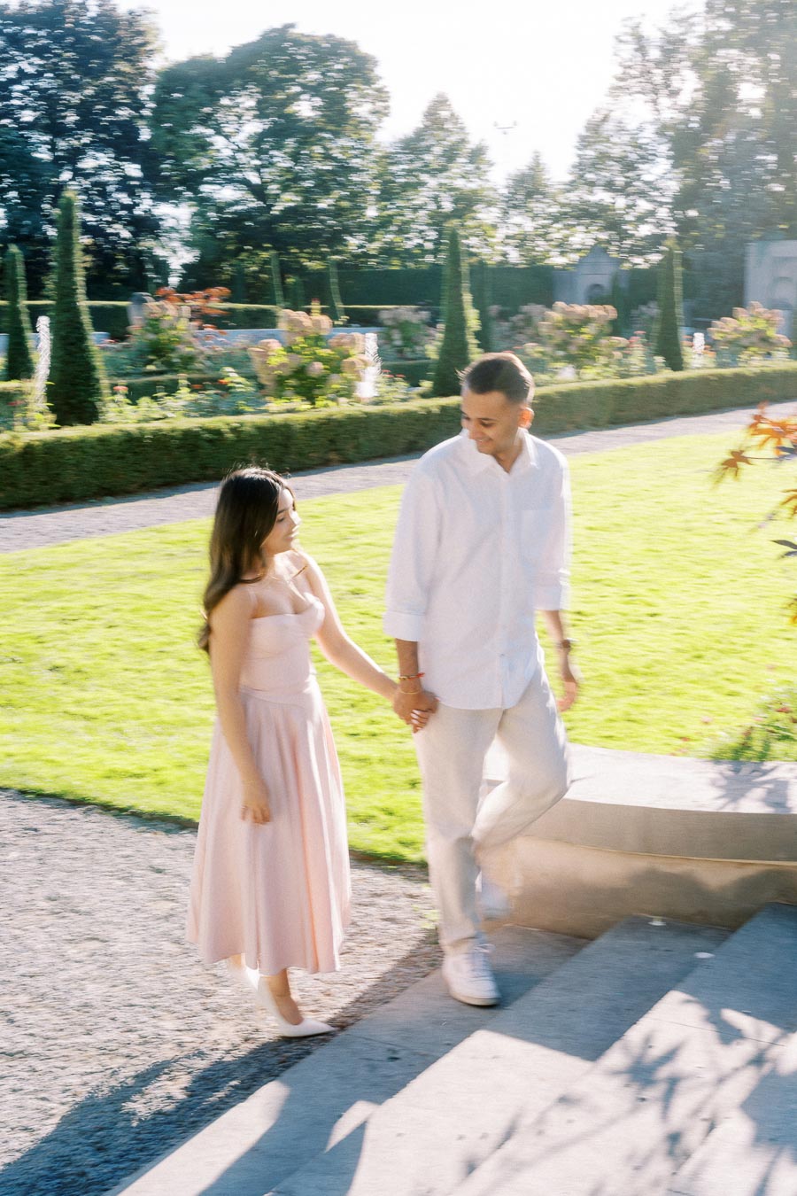 A couple holding hands and walking up stone steps in a vibrant garden setting with manicured bushes and sunny weather.