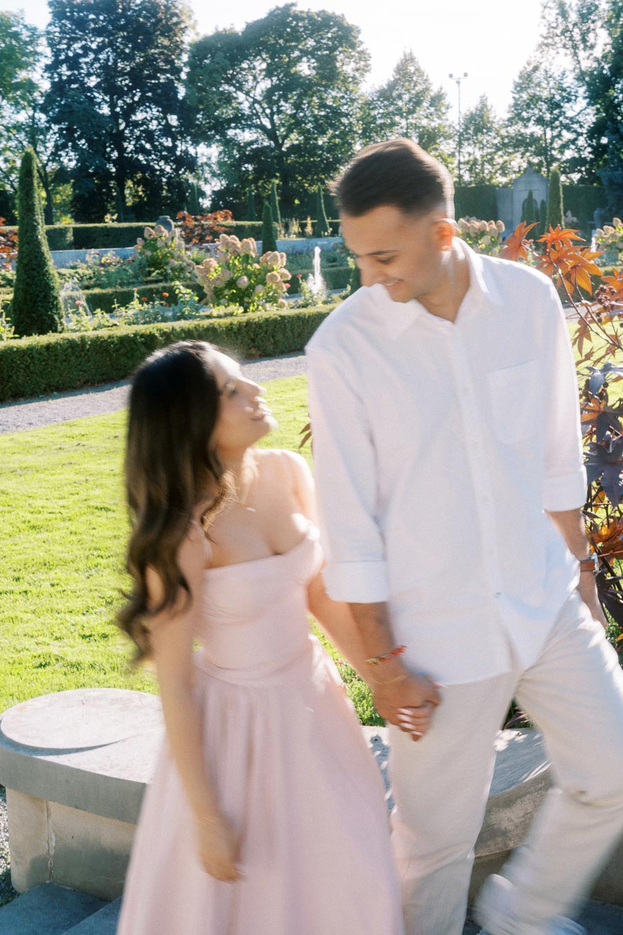 A couple smiling and holding hands in a lush green garden during a sunny day, surrounded by flowers and trees.