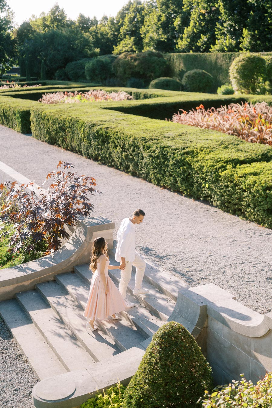 A couple holding hands while walking up stone steps in a beautifully manicured garden with lush hedges and vibrant plants under a clear blue sky.