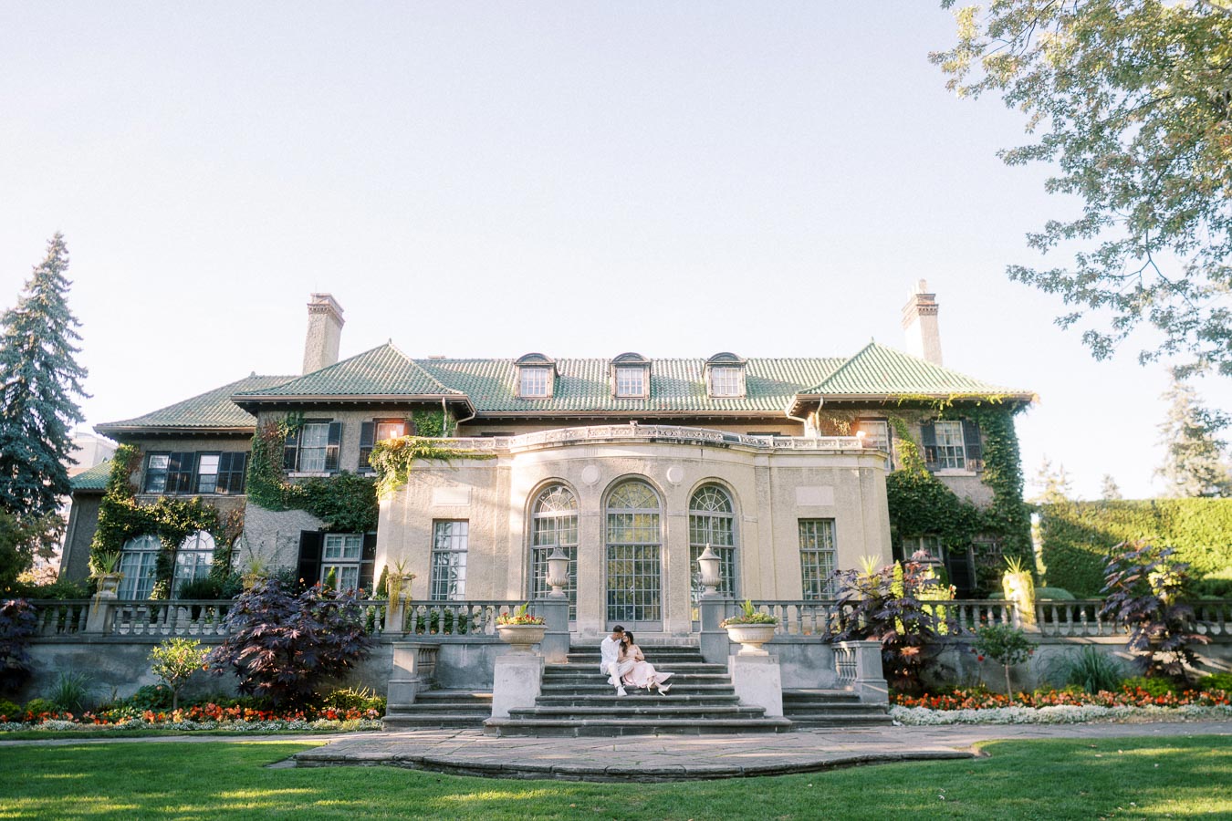 Elegant historic mansion with ivy-covered walls and lush gardens, featuring a couple sitting on the stone steps under clear blue skies.