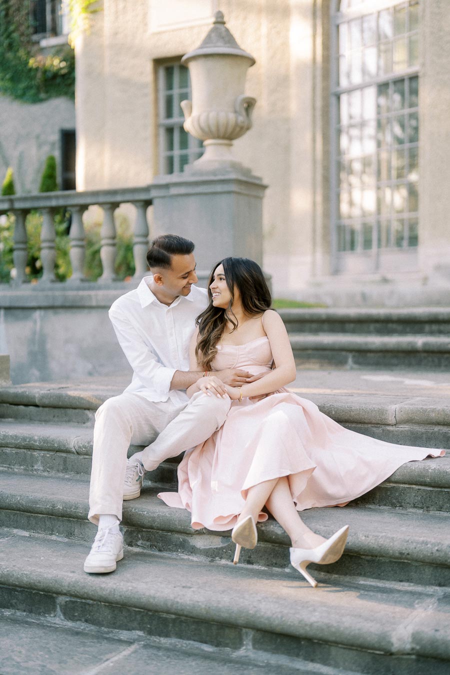 A couple sitting lovingly on stone steps in front of an elegant building, wearing light pastel outfits, with greenery in the background, conveying romance and elegance.