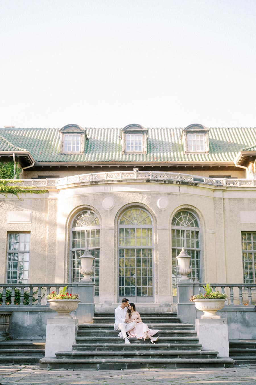 A couple sitting on elegant stone steps in front of a historic building with large arched windows and a green tiled roof, surrounded by decorative planters.