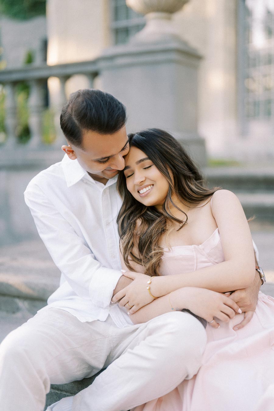 A young couple in love embracing outdoors, dressed in elegant attire with soft smiles, sitting on steps in a romantic setting.