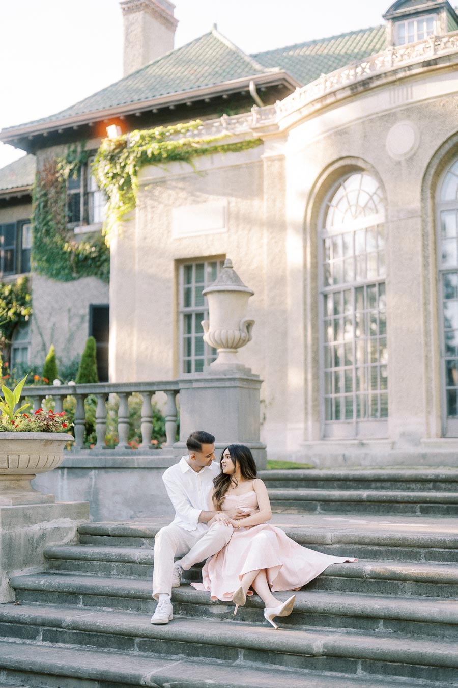 A couple sitting on stone steps outside a historic building, surrounded by lush greenery and elegant architecture, creating a romantic atmosphere.