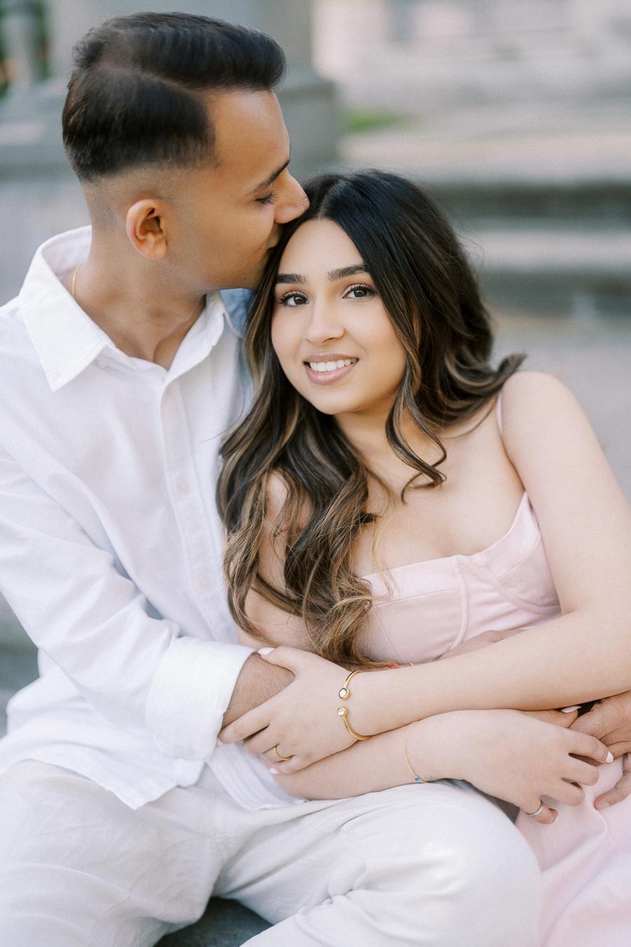 Couple sitting and embracing on stone steps, with the man kissing the woman's forehead. They both appear content and relaxed, wearing light-colored clothing, enhancing the warm and romantic atmosphere.