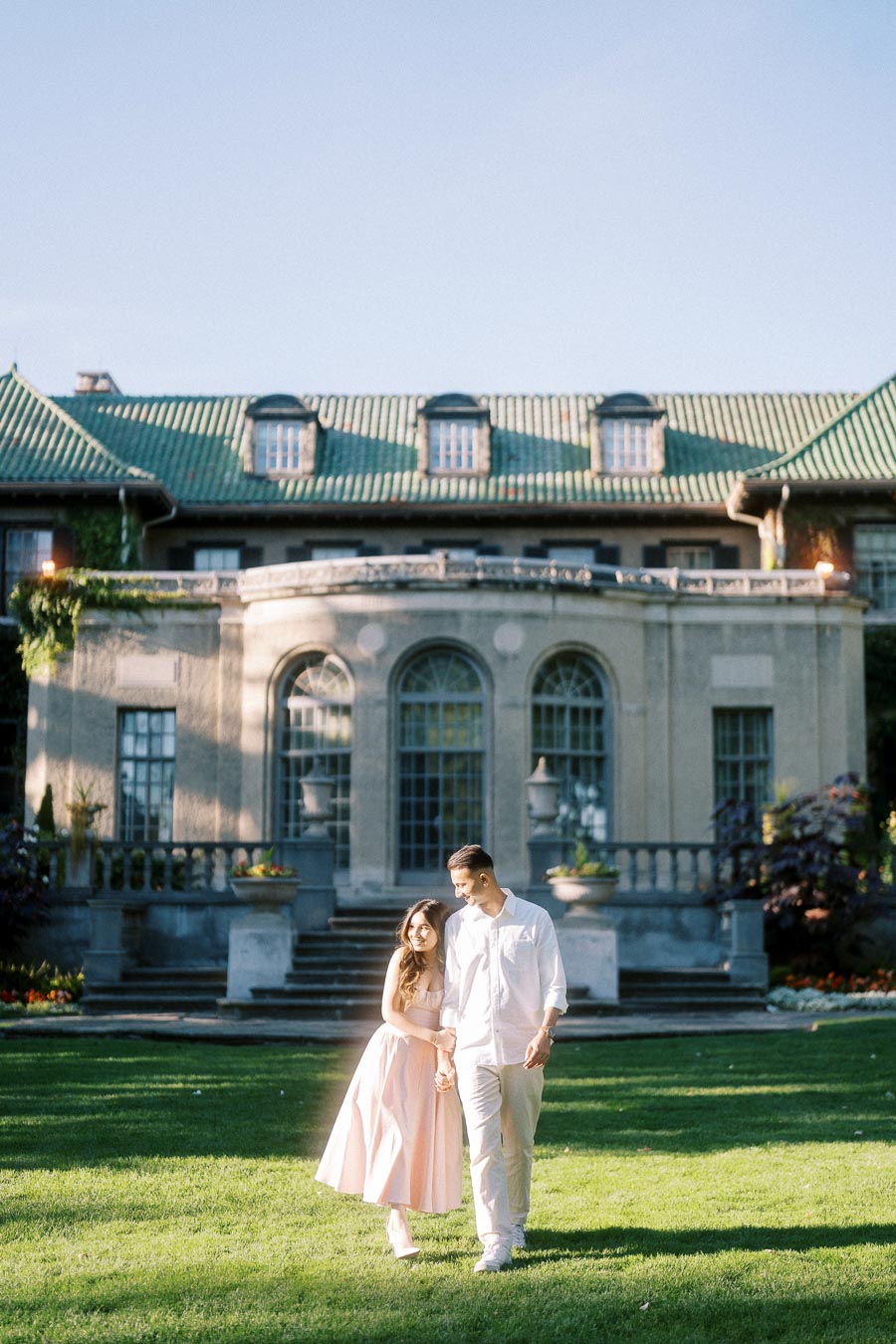 Romantic couple walking hand in hand in front of a historic mansion with lush green lawn and elegant architecture under a clear blue sky.