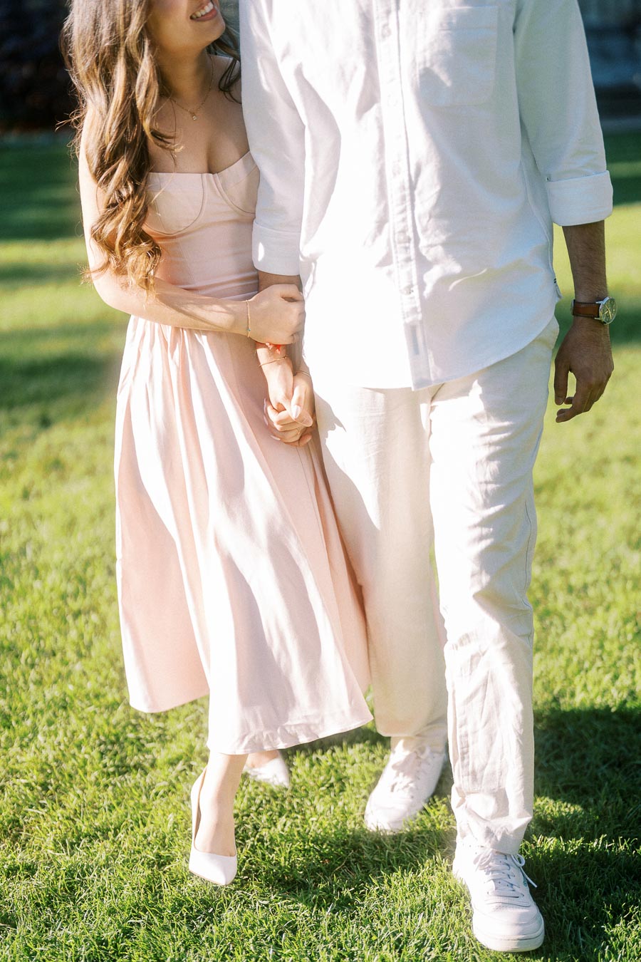 A couple walking hand in hand on a sunny day in a garden, wearing light-colored attire.
