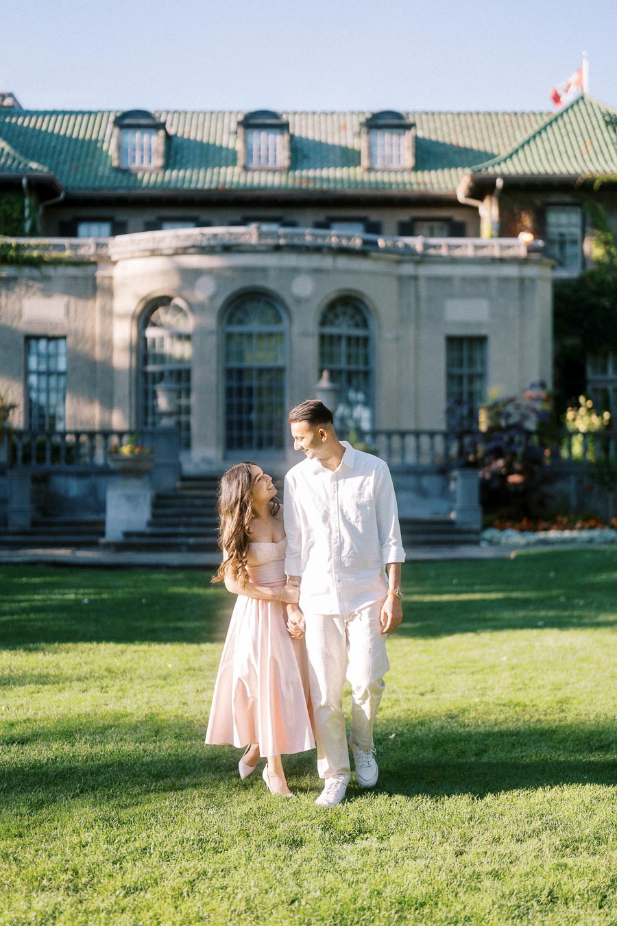 A couple walking hand in hand on a sunny day in front of a historic building with a manicured lawn. The woman is wearing a pink dress, and the man is dressed in white, both smiling and enjoying the moment.