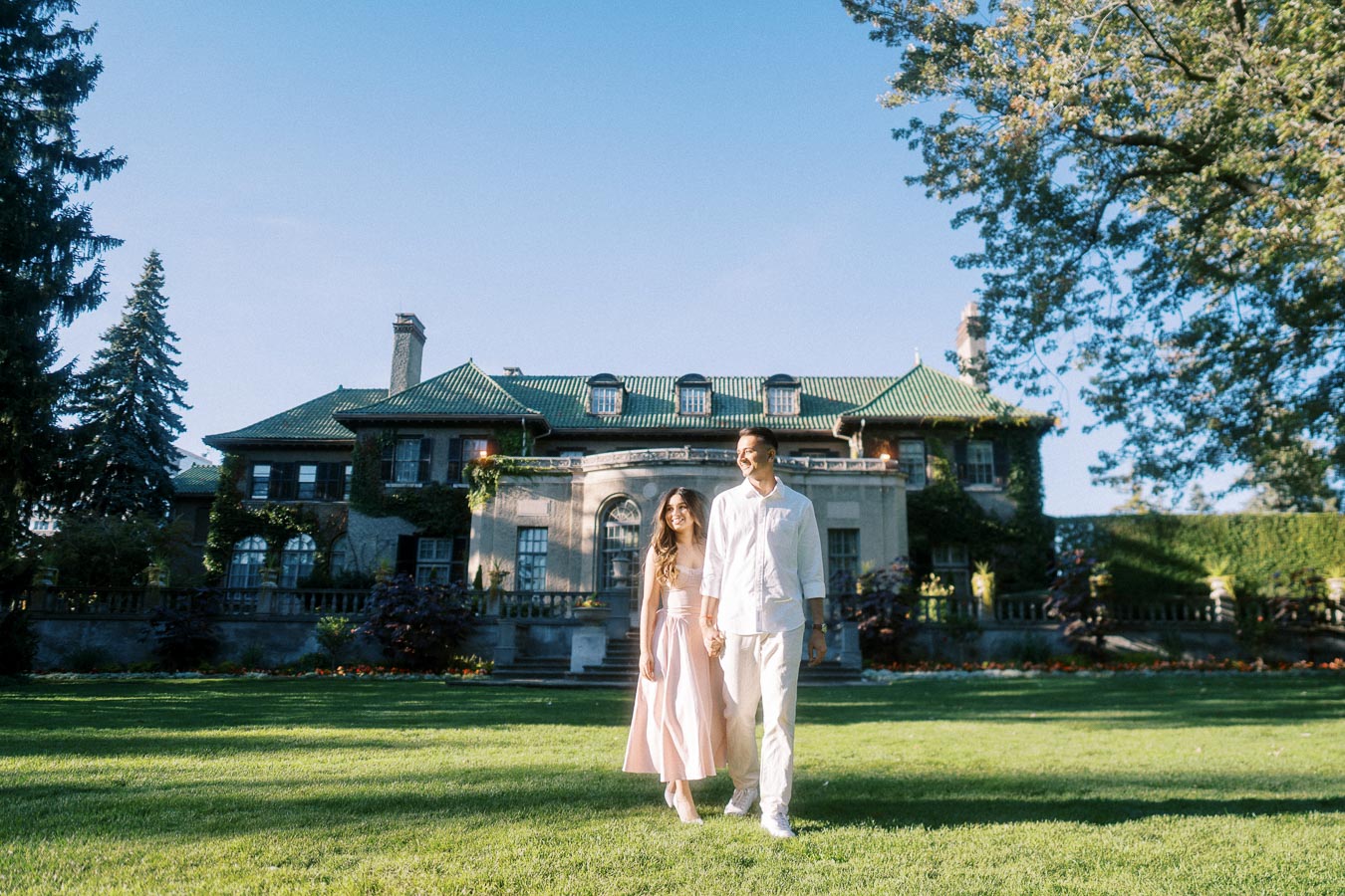 A couple walking hand in hand in front of a historic mansion, surrounded by lush greenery on a sunny day.