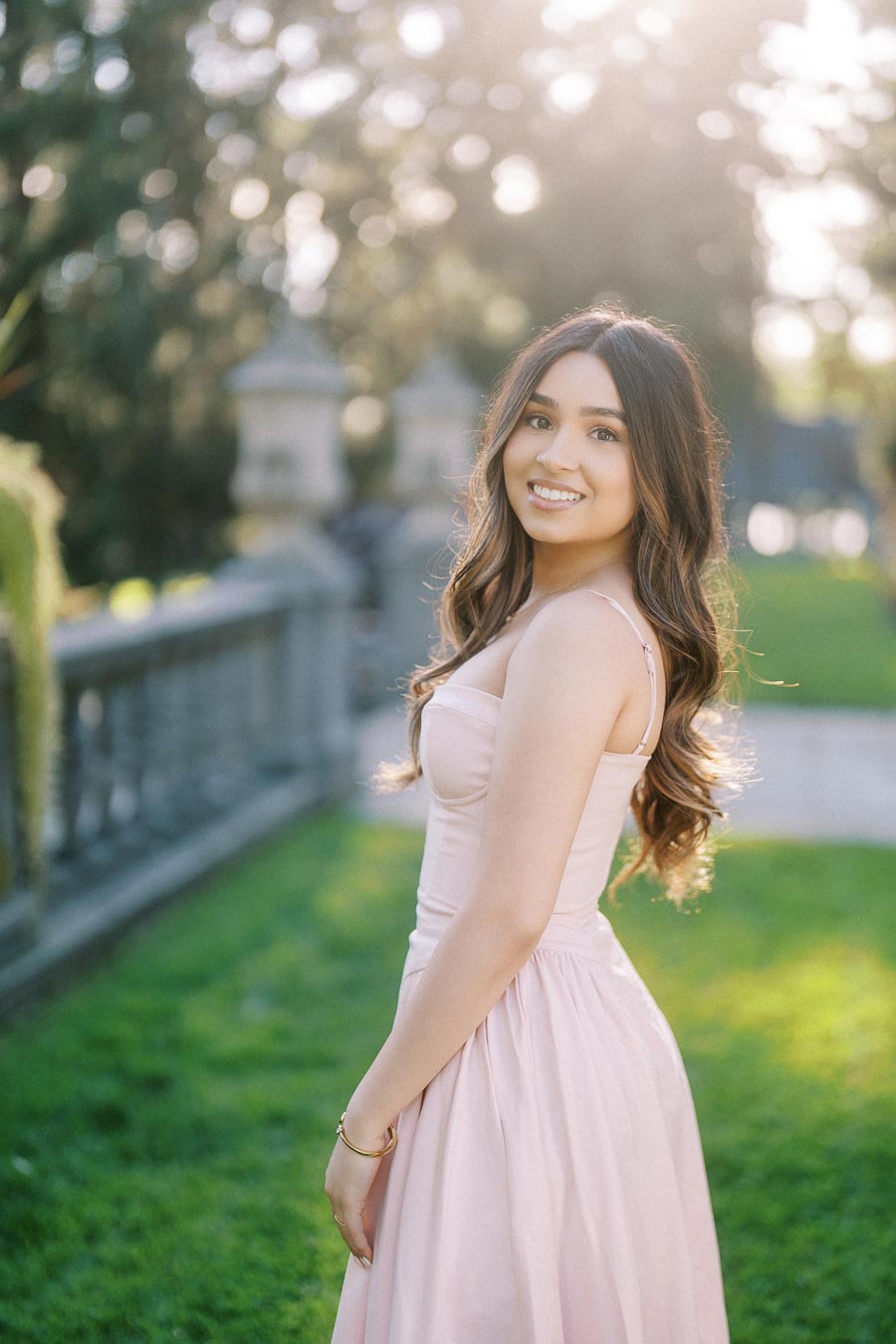 Young woman in a light pink dress smiling in a sunlit garden setting, surrounded by lush greenery and a stone balustrade.