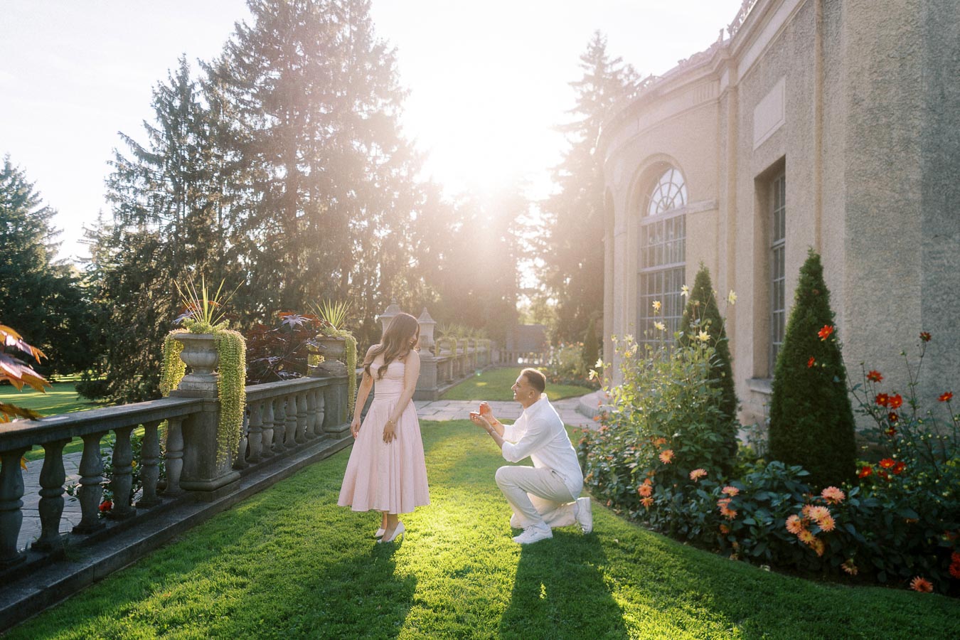 A man proposes to a woman in a sunny garden setting near an elegant building, surrounded by lush greenery and colorful flowers.