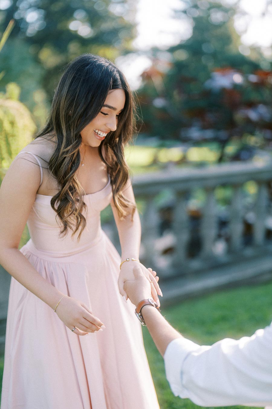 Young woman in a pink dress smiling and holding hands with another person, set in a sunny garden with greenery in the background.