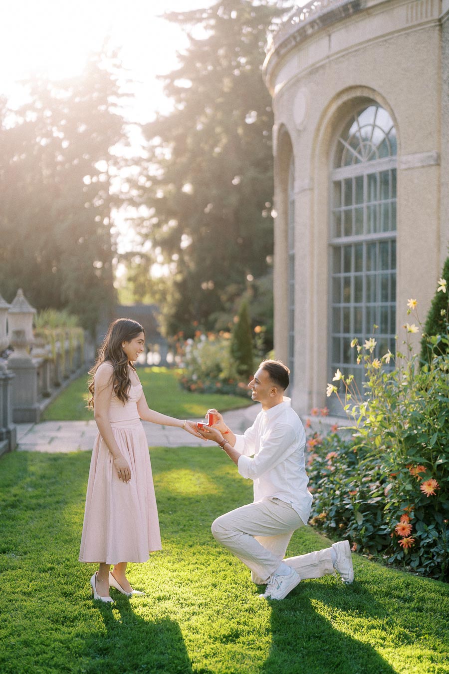 A romantic proposal scene in a garden with a man kneeling and holding a small red box towards a woman in a light pink dress, surrounded by lush greenery and elegant architecture.
