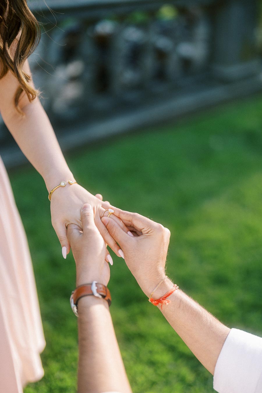 A person kneeling and placing a gold engagement ring on another person's finger, set against a lush green garden background.