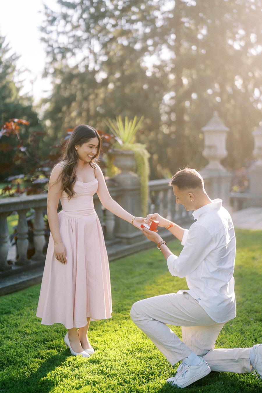 A man kneels on a grassy lawn in a romantic garden setting, proposing to a smiling woman in a pink dress.