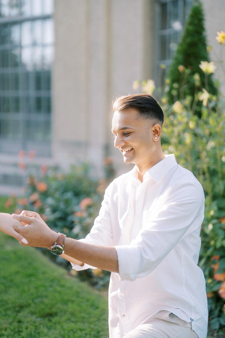 A man in a white shirt smiling and holding hands with someone outdoors, surrounded by greenery and flowers, in a sunlit garden setting.