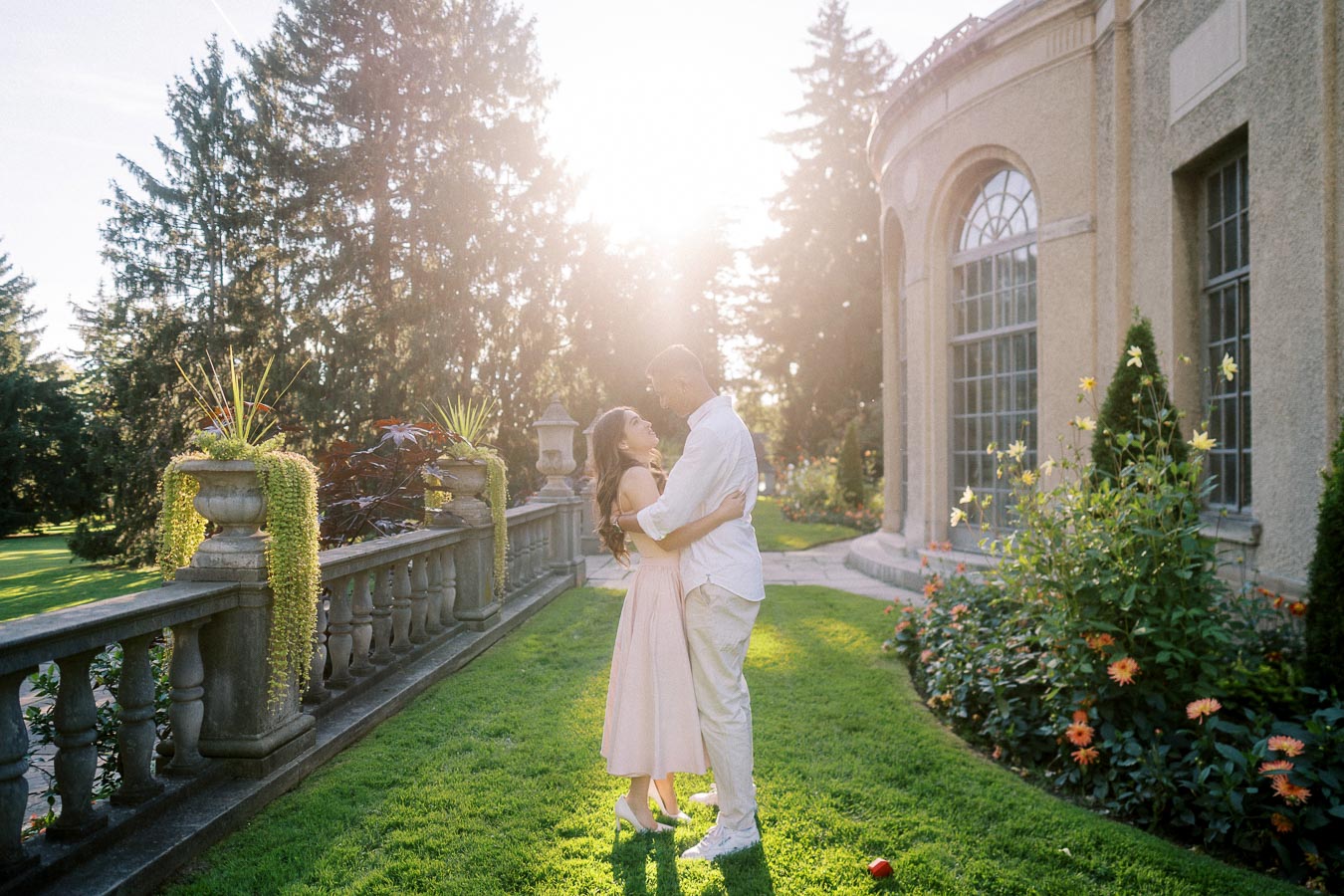 A couple embracing in a sunlit garden beside a historic building, surrounded by lush greenery and colorful flowers, creating a romantic atmosphere.