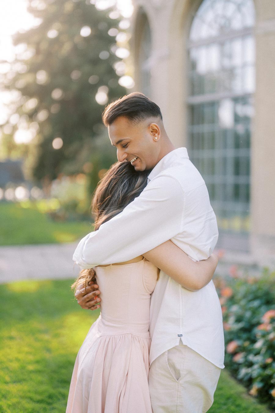 A couple embraces lovingly in a sunlit garden, with blooming flowers and a vintage building in the background. The man, wearing a white shirt, gently holds the woman, who is dressed in a light pink dress, showcasing a moment of affection and happiness.