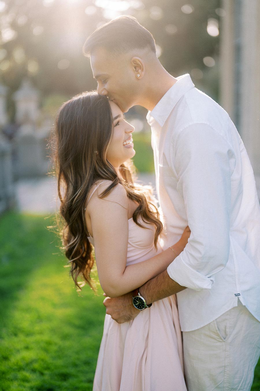 A couple embracing in a sunlit garden, with the man gently kissing the woman's head. Both are smiling and dressed in light-colored clothing, surrounded by lush greenery and a soft, blurred background.