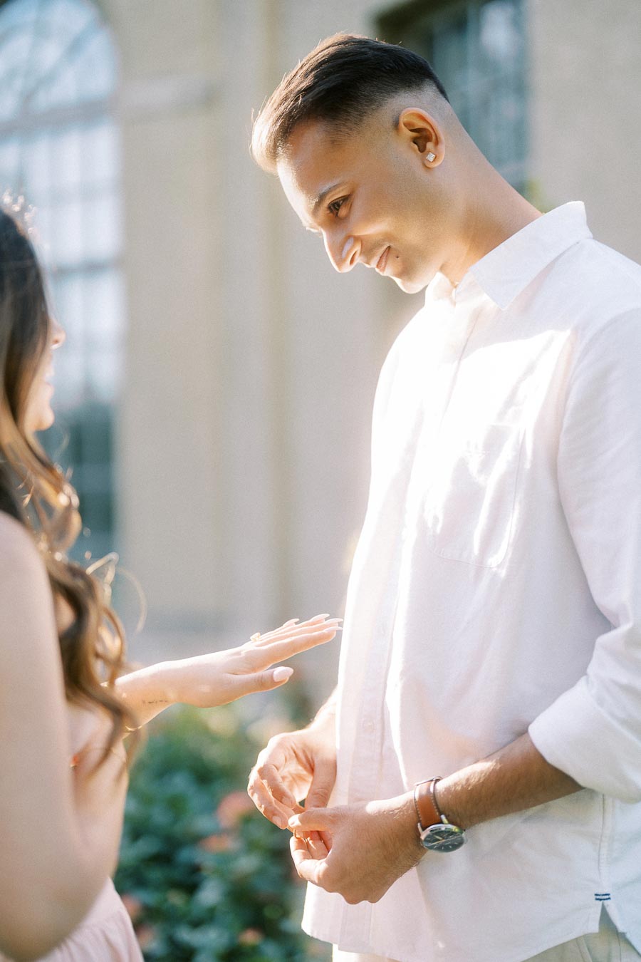 A man in a white shirt places a ring on a woman's finger during a sunlit engagement moment, with a garden setting in the background.