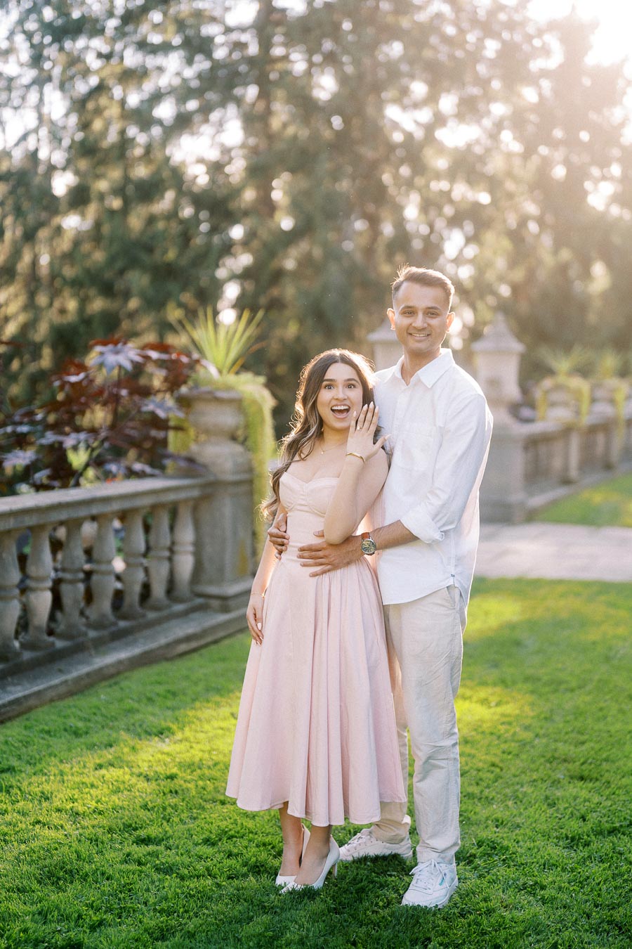 A happy couple celebrating their engagement outdoors, with the woman in a pastel pink dress showcasing her engagement ring, while the man lovingly embraces her from behind in a lush garden setting with sunlight filtering through the trees.
