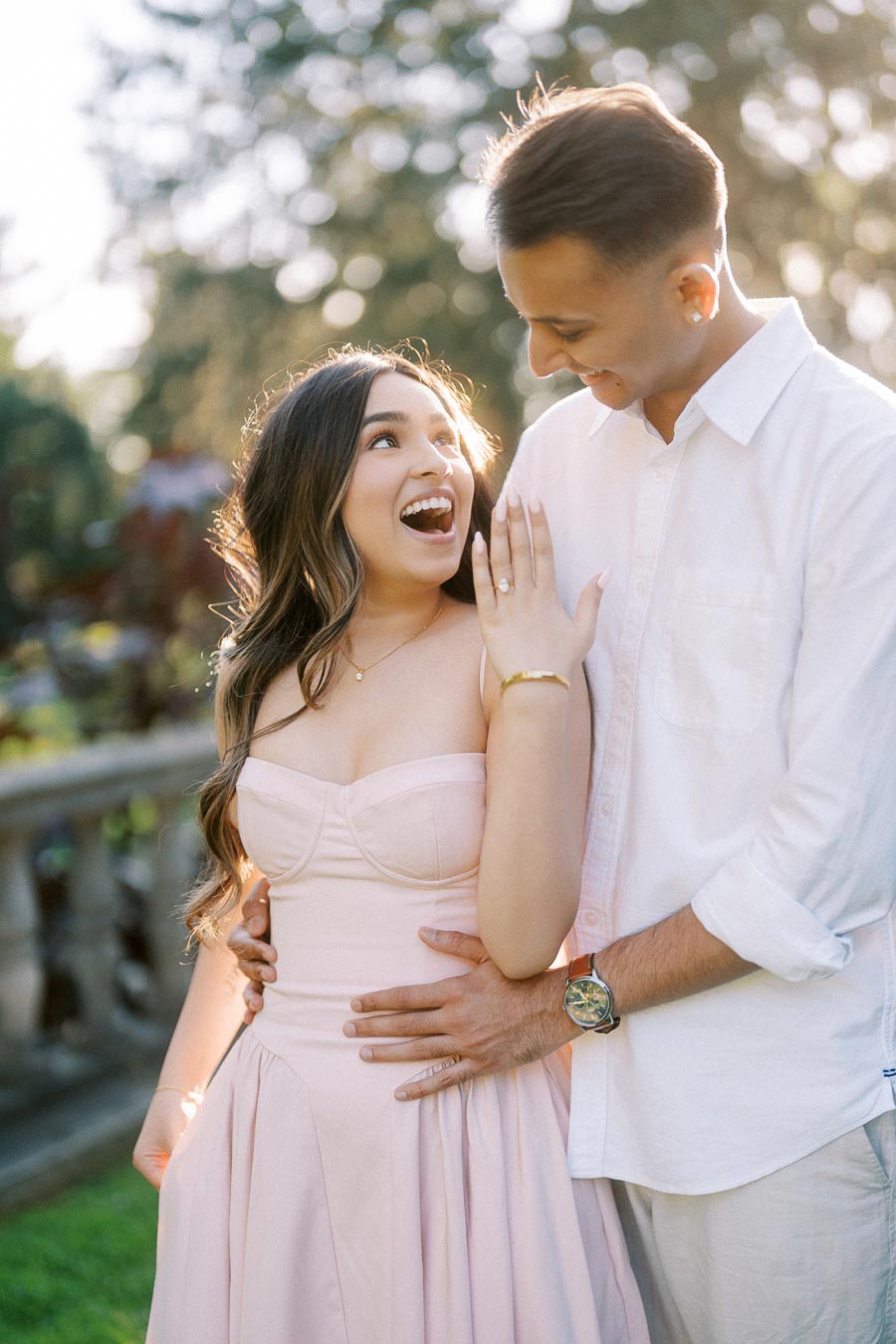 Happy couple celebrating engagement, woman showing engagement ring while embraced by her partner in a sunlit garden setting.