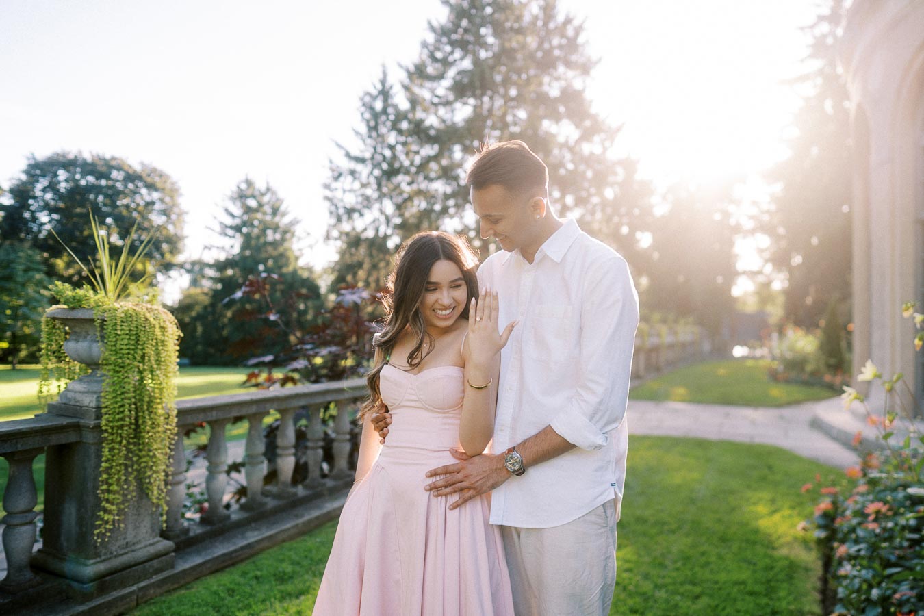 A happy couple celebrating their engagement in a sunny garden, with the woman showing her engagement ring. The man lovingly stands beside her, both dressed in elegant pastel attire, surrounded by lush greenery and floral decorations.