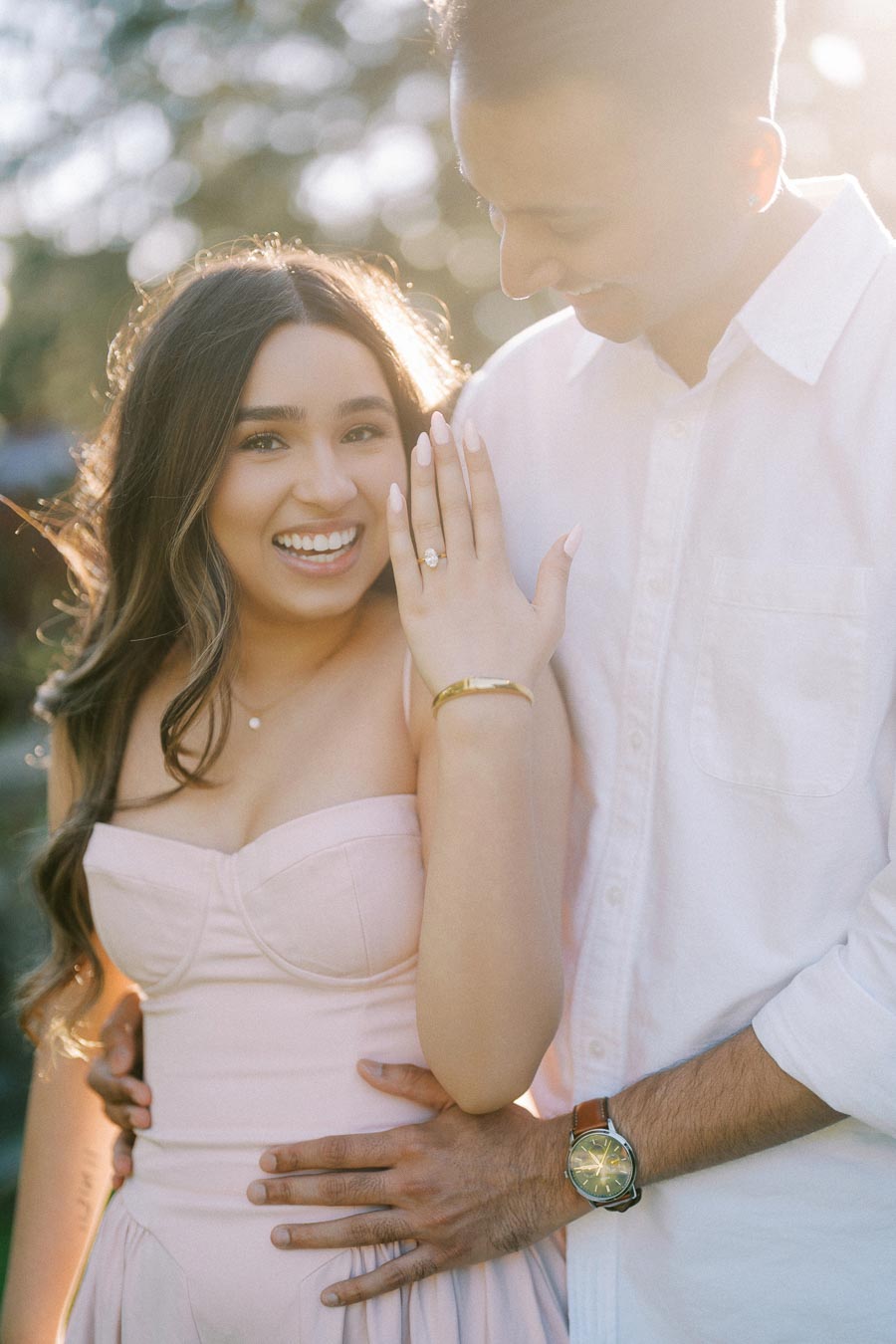 Happy engaged couple in a garden setting, woman showing her engagement ring with a joyful smile and sunlight in the background.