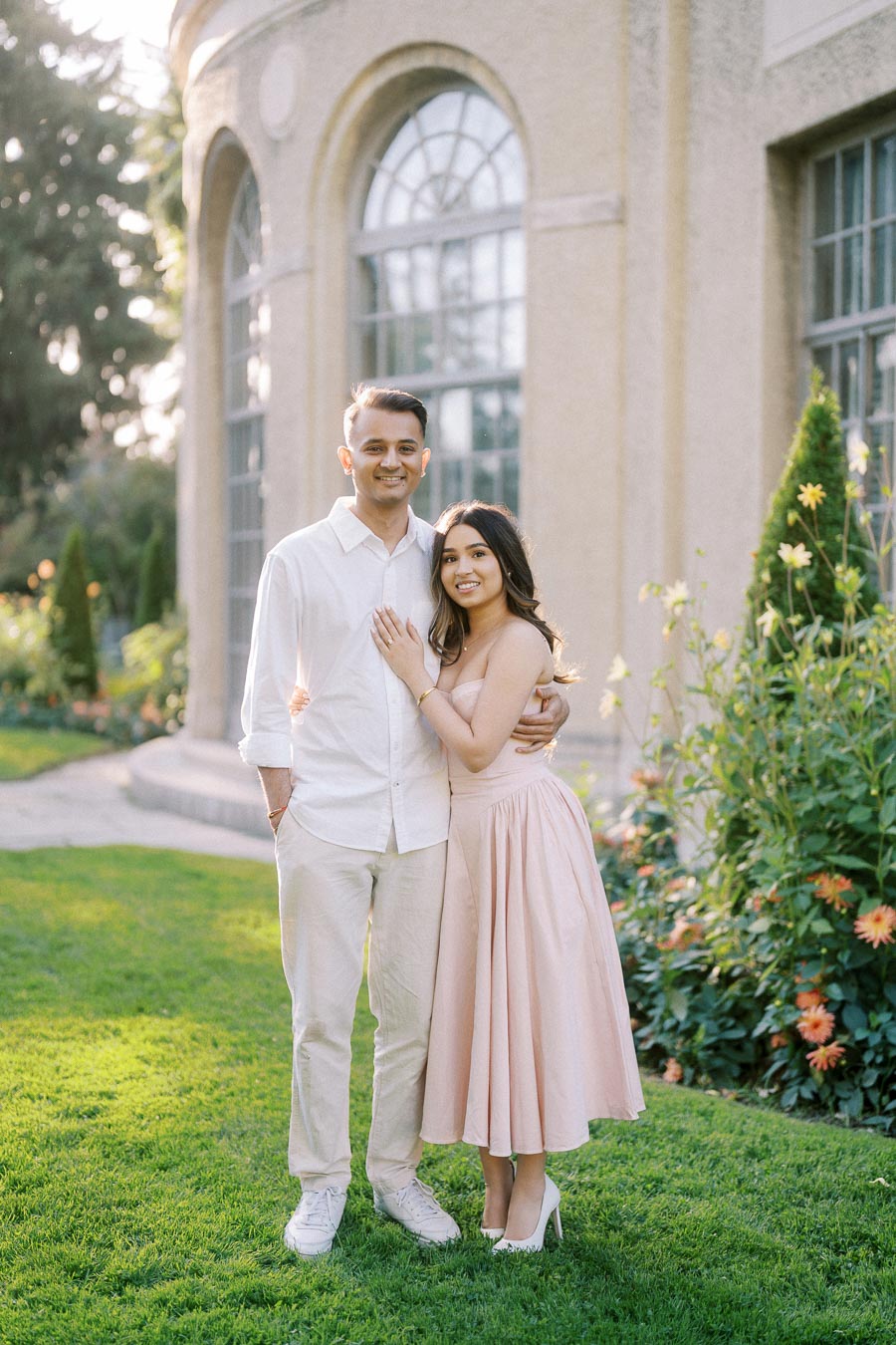 Couple posing together in a garden setting with a historic building in the background, dressed in light pastel attire, surrounded by greenery and blooming flowers.