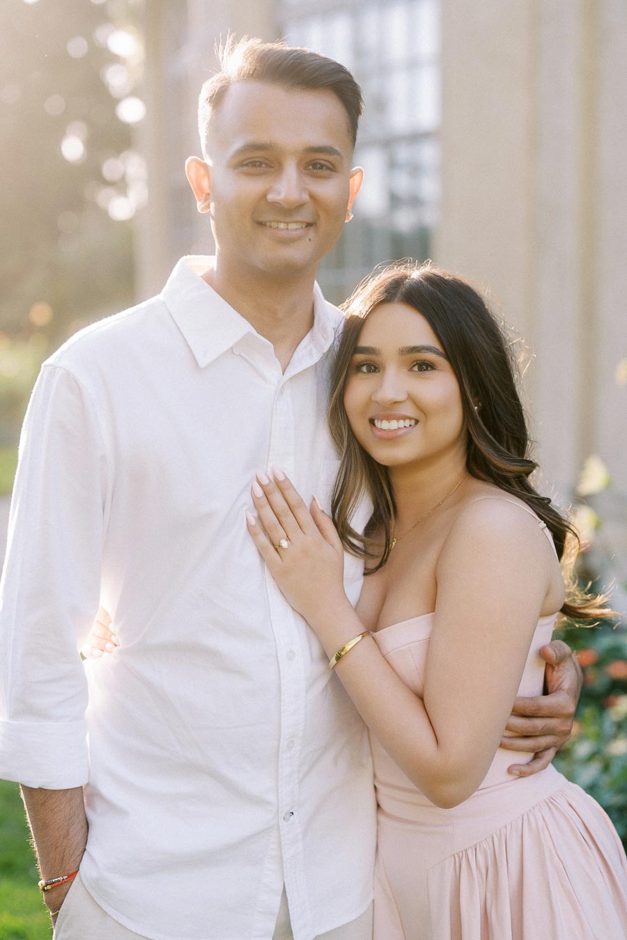 Engaged couple smiling in a sunlit garden, the woman wearing a strapless pink dress and displaying an engagement ring, while the man wears a white shirt, creating a romantic and joyful scene.