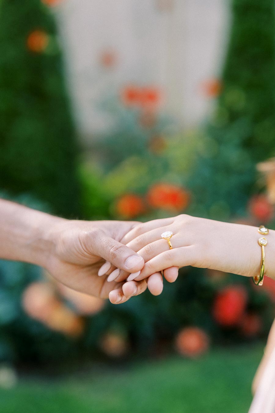 Two people holding hands with one wearing a diamond engagement ring and gold bracelet, set against a blurred garden background with colorful flowers.