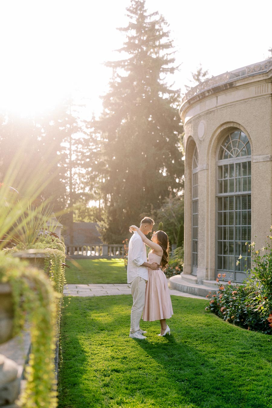 A couple embracing on a sunlit lawn in front of an elegant building with arched windows, surrounded by lush greenery and trees.