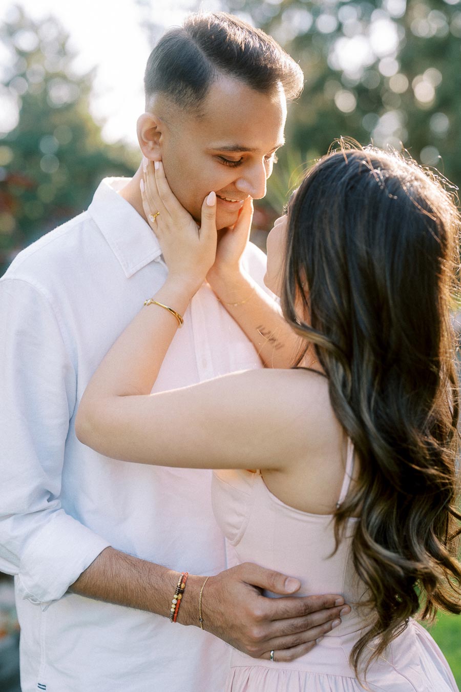 A couple embracing and smiling, sharing an intimate moment outdoors with soft natural lighting and blurred greenery in the background.