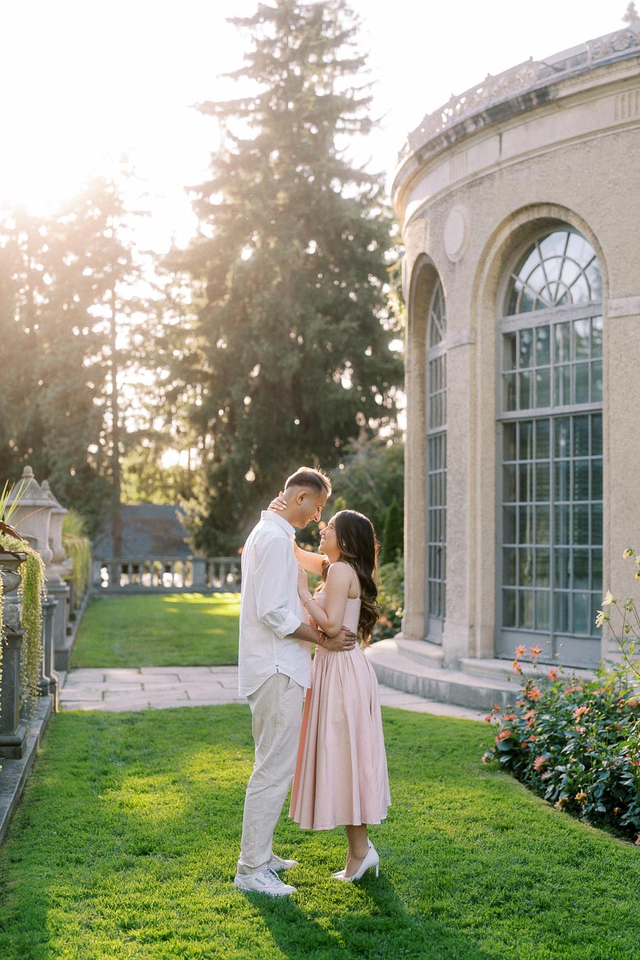 A couple sharing a romantic moment in a garden setting, with the sun shining through tall trees in the background and a historic building with arched windows nearby.