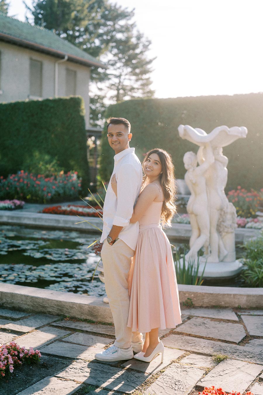 A couple posing joyfully in a garden setting with a pond and statue, dressed in light pastel colors, surrounded by lush greenery and vibrant flowers under a clear sky.