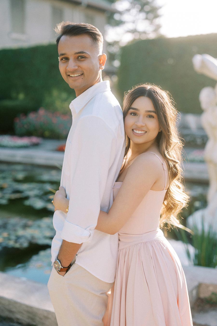 Happy couple posing in a garden setting, with the woman in a pastel pink dress hugging the man in a white shirt, surrounded by greenery and a pond.