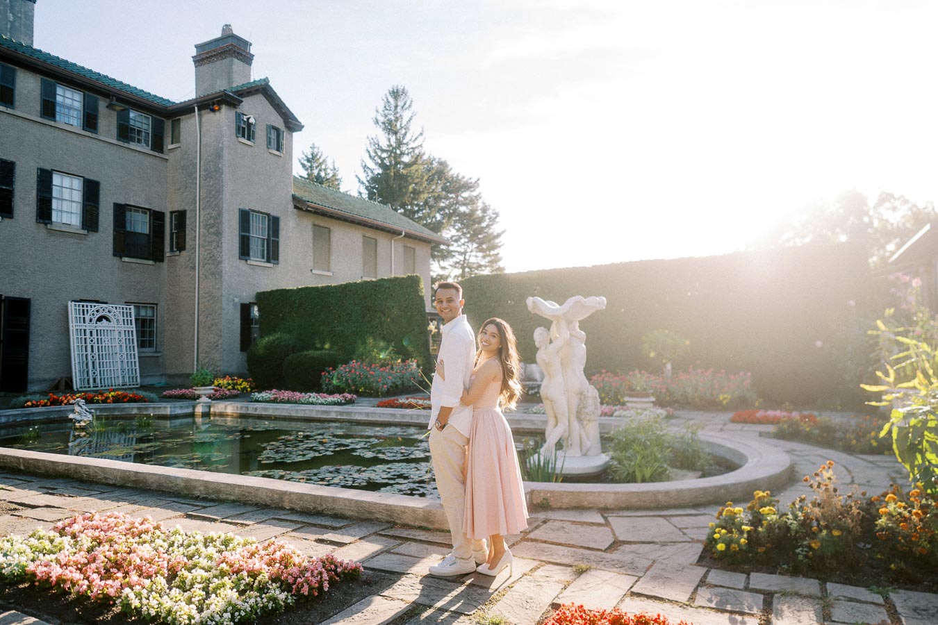 A happy couple posing in front of a historic mansion, surrounded by a beautiful garden with vibrant flowers and a decorative pond, under the warm glow of sunset.
