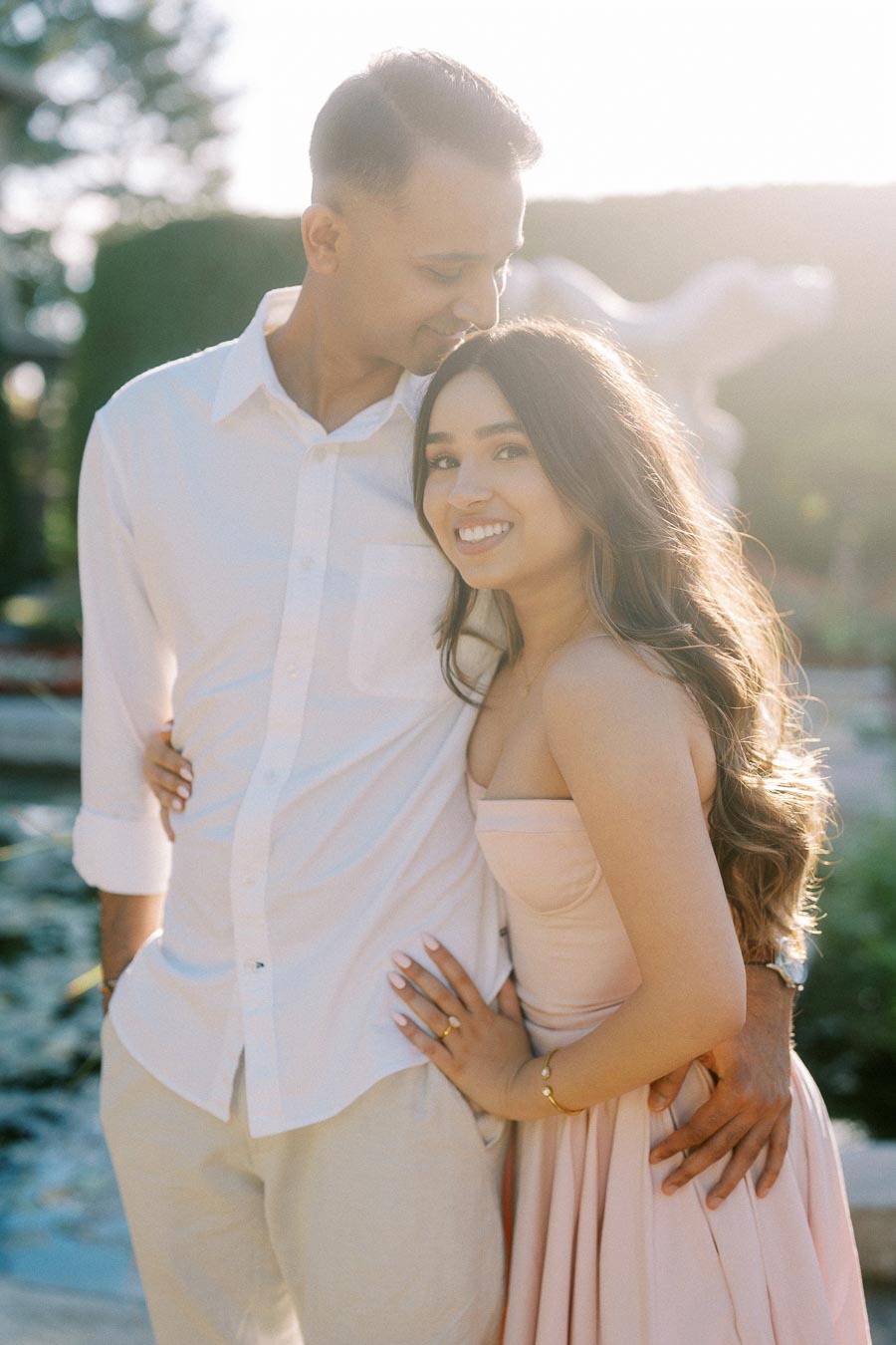 A couple embracing and smiling in a sunlit garden setting, wearing light-colored outfits, with greenery and soft sunlight in the background.