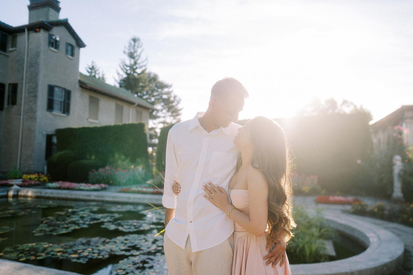 A couple embraces lovingly in a sunlit garden setting with a pond and historic building in the background, capturing a romantic moment.
