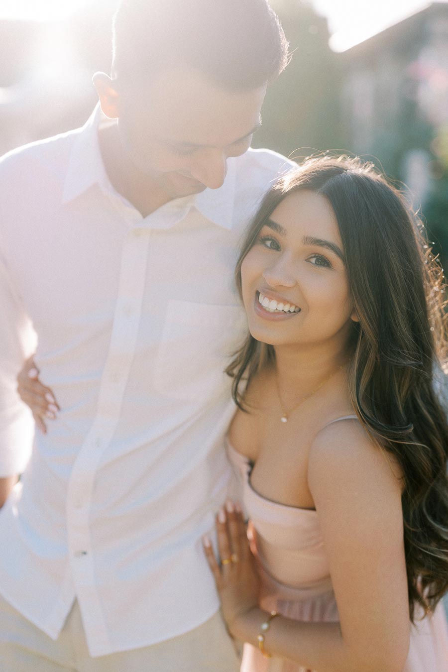 Smiling couple in sunlight, with woman in a pink dress and man in a white shirt embracing outdoors.