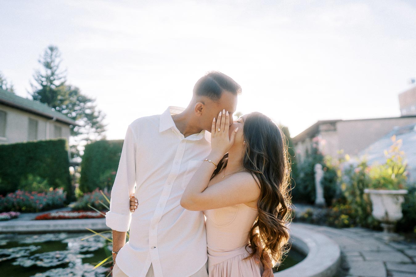 A couple sharing a romantic kiss by a garden fountain, with sunlight illuminating their embrace and surrounding greenery.