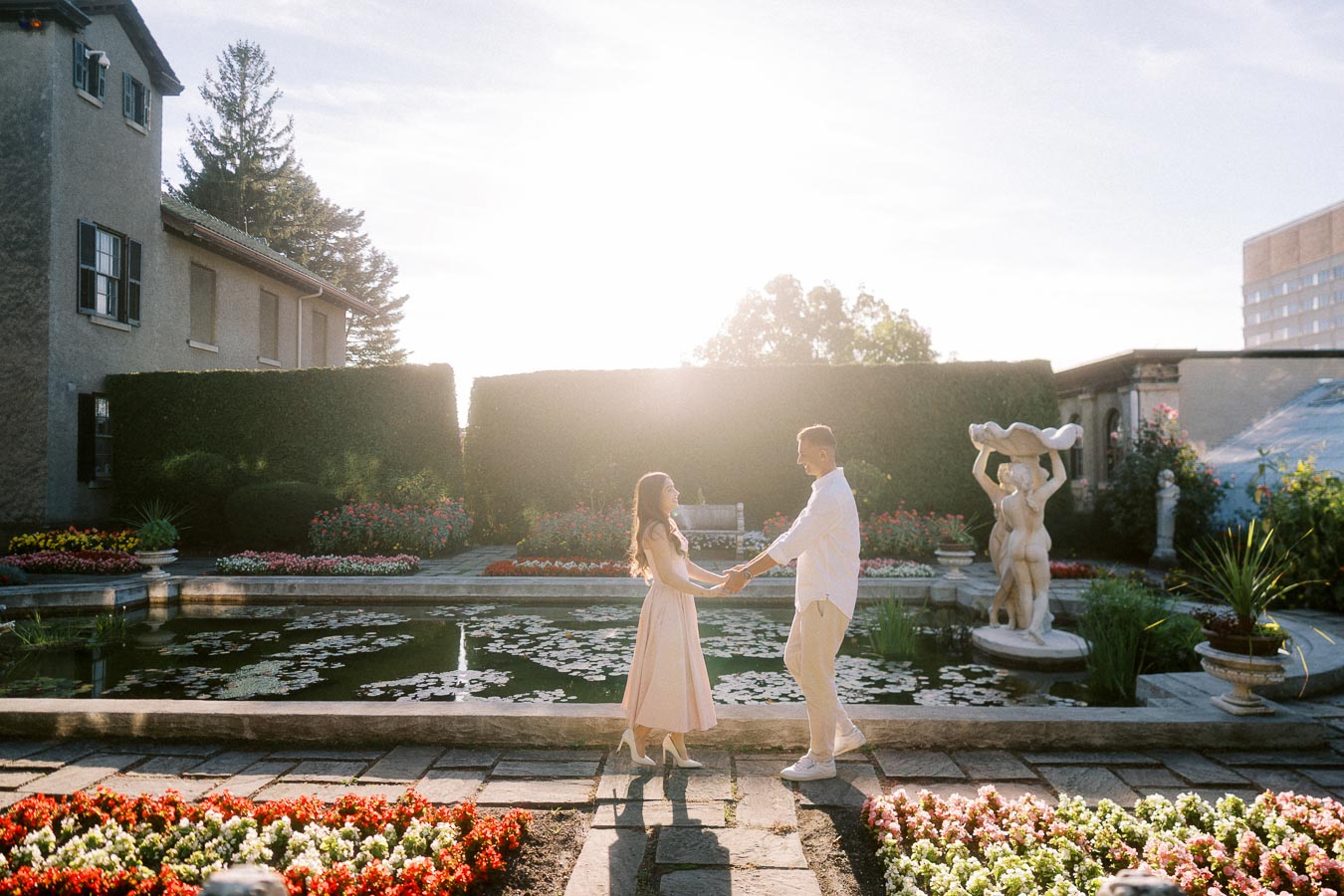 A couple holding hands in a beautifully landscaped garden at sunset, with a fountain, colorful flower beds, and a statue in the background.