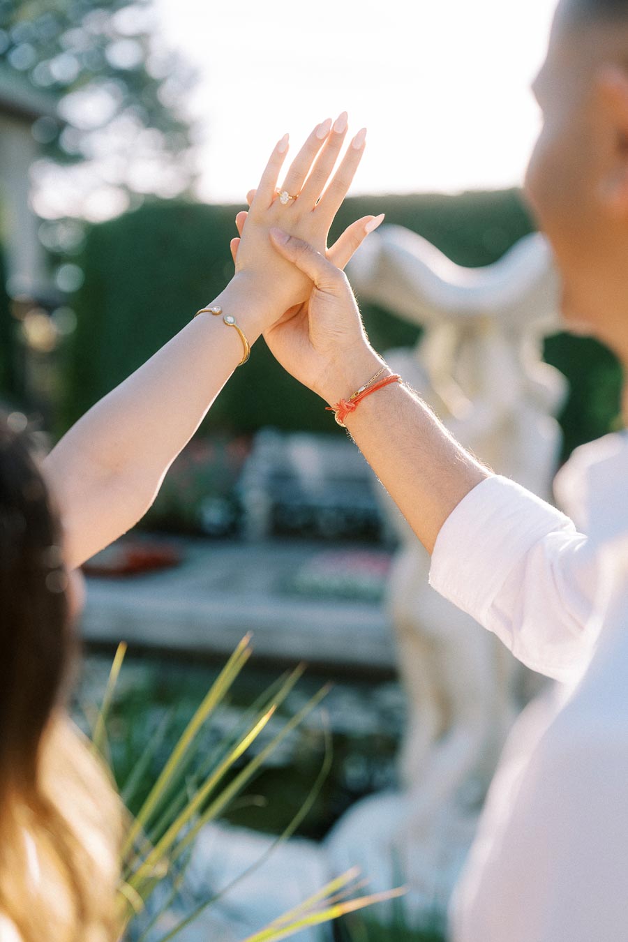 A couple joyfully holding hands, showing off elegant jewelry against a serene garden backdrop, highlighting love and commitment.