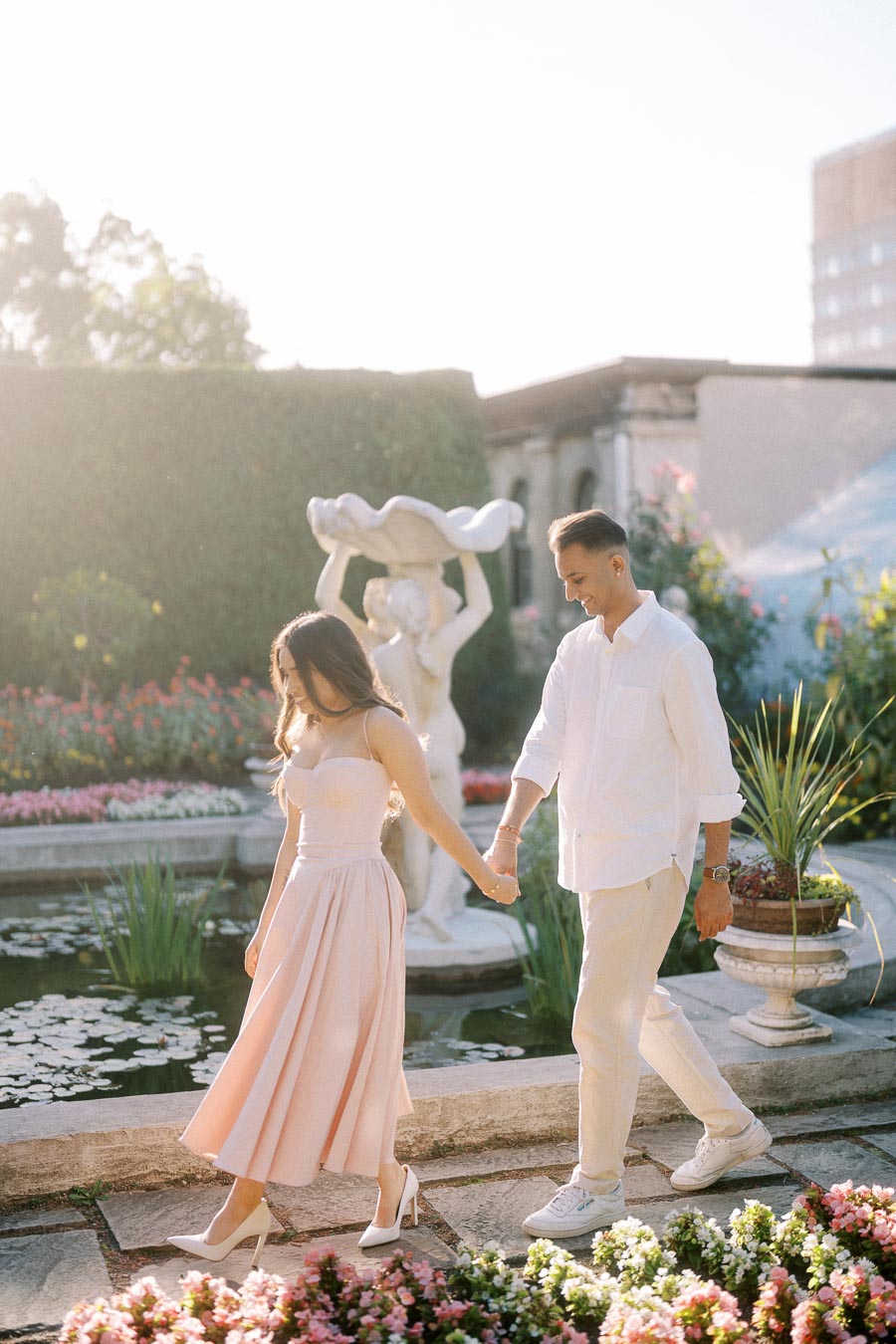 A couple holding hands walks beside a garden pond with blooming flowers and a decorative statue in the background, illuminated by soft sunlight.