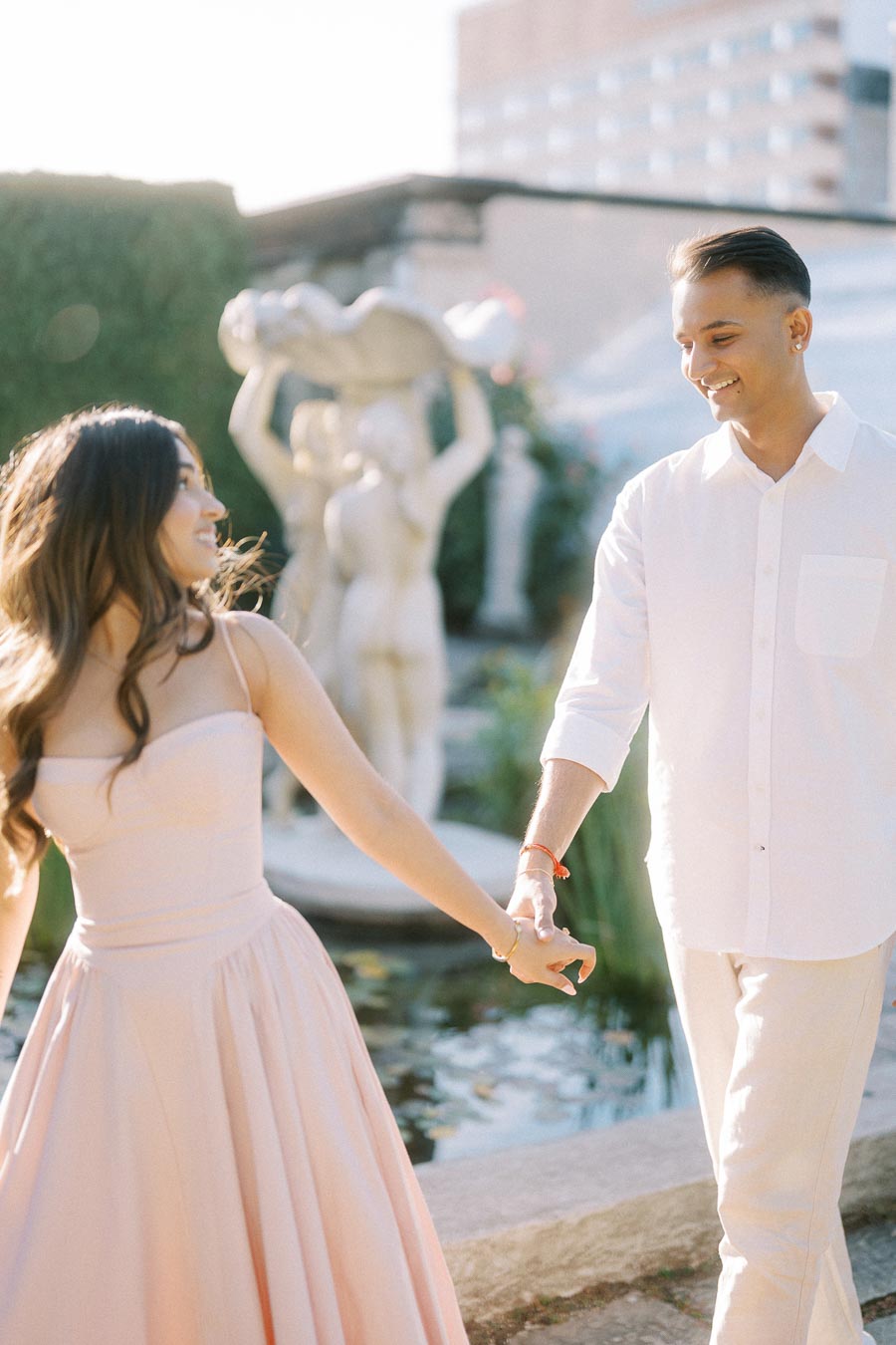Couple holding hands in a garden setting, dressed in pastel attire, with a sculpture and building in the background on a sunny day.