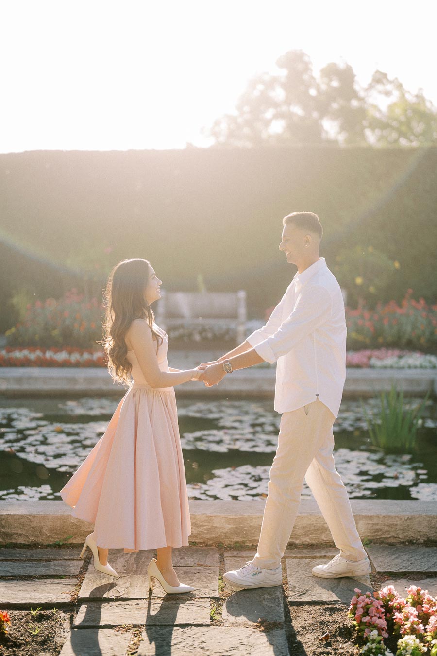 A couple holding hands and smiling at each other in a sunlit garden next to a lily pond, surrounded by blooming flowers.