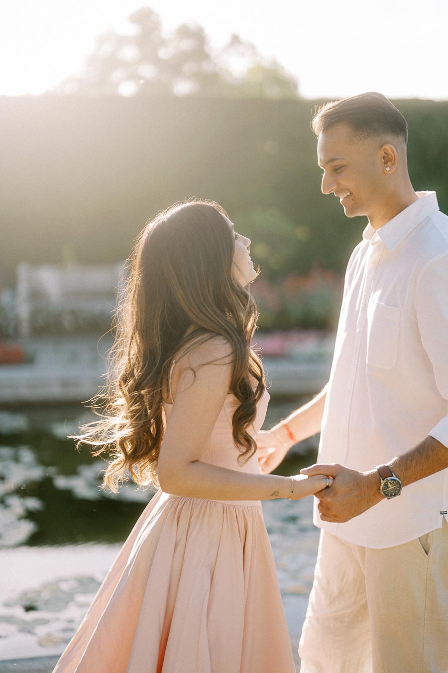 A couple sharing a joyful moment in a sunlit garden, holding hands with smiles, surrounded by lush greenery and a serene pond, conveying a feeling of romance and happiness.